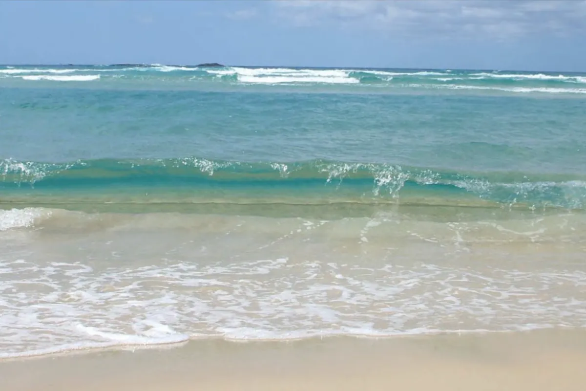 View from the water's edge of a surf beach, with a calm turquoise gutter in the middle and a line of waves breaking over the outer sandbank near the horizon