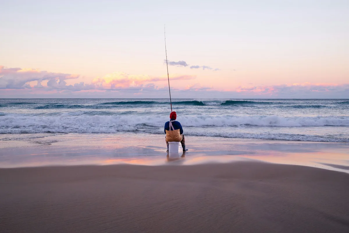 Surf angler sitting on a bucket at the water's edge at dawn, rod up, with waves breaking on an Australian beach